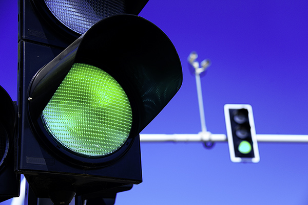 Nahaufnahme einer Grünen Ampel mit einer weiteren grünen Ampel im Hintergrund, blauer Himmel